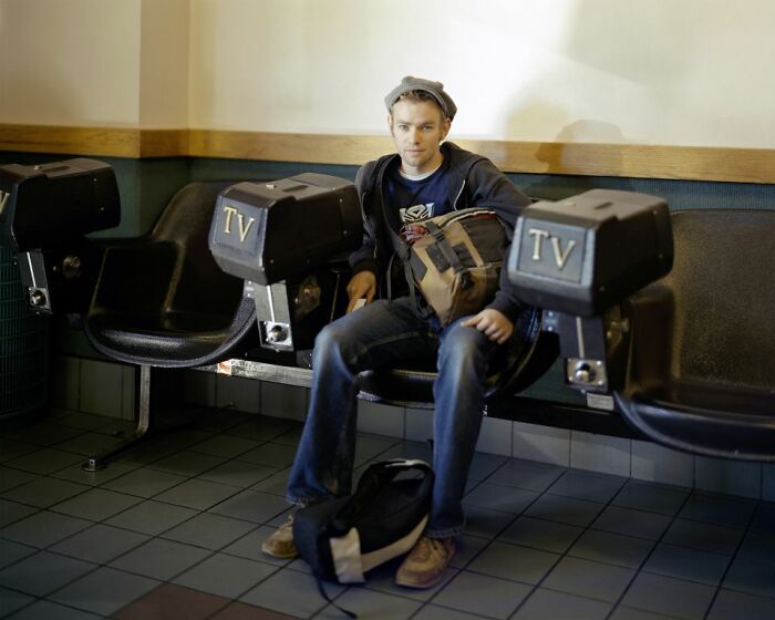 Young man with a backpack sitting at a bus stop with old TV sets attached to the seats, documenting life in America.