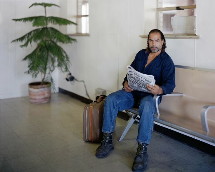Man sitting at a bus stop bench reading a newspaper, with a suitcase beside him, documenting life at America’s bus stops.