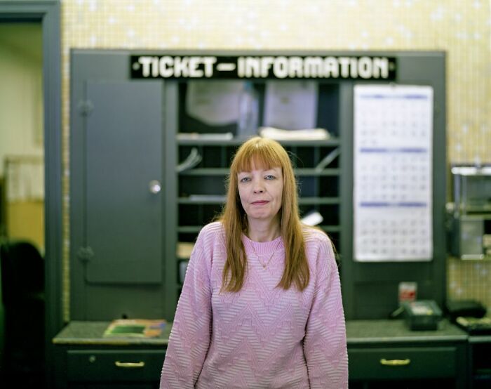 Woman in a pink sweater stands at a ticket information booth, capturing life at America’s bus stops in a photo series.