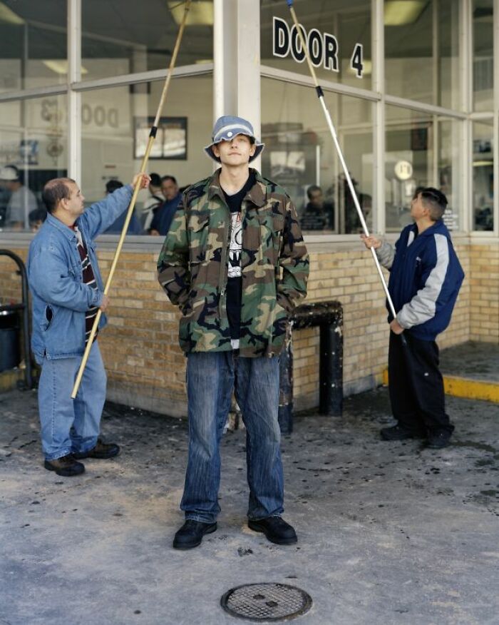 Young man in camo jacket and bucket hat stands outside a bus stop while others clean windows behind him.