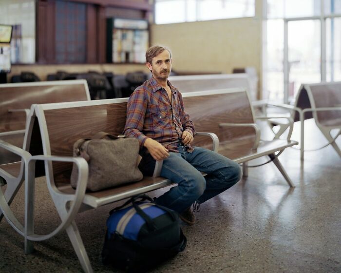 Man in plaid shirt sitting with bags on bench at a bus stop, capturing life in America by bus photography.