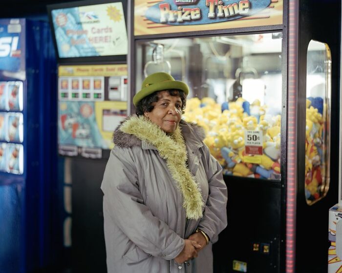 Elderly woman in a green hat and coat standing by arcade claw machine capturing life at America’s bus stops.