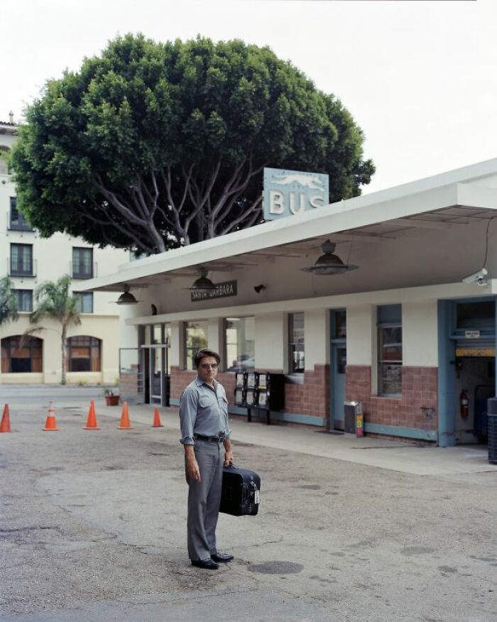 Man standing at an America bus stop holding a suitcase under a large tree near a vintage bus station building.
