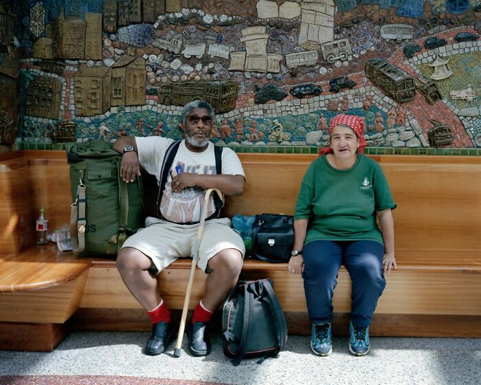 Two people sitting at a bus stop with bags, captured by a photographer documenting life at America's bus stops.