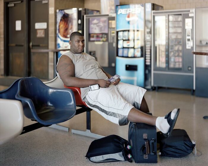 Man sitting at a bus stop with luggage, wearing shorts and sandals, captured in a life at America’s bus stops photo.