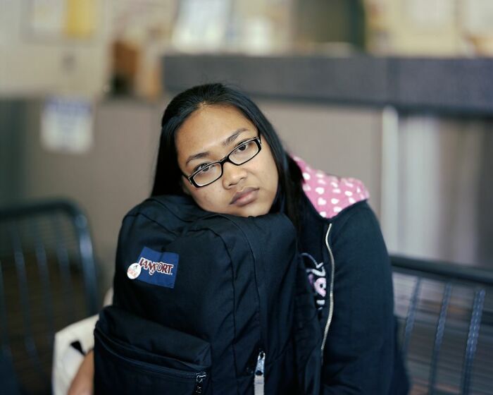 Woman wearing glasses resting head on black backpack while waiting at a bus stop, capturing life at America’s bus stops.