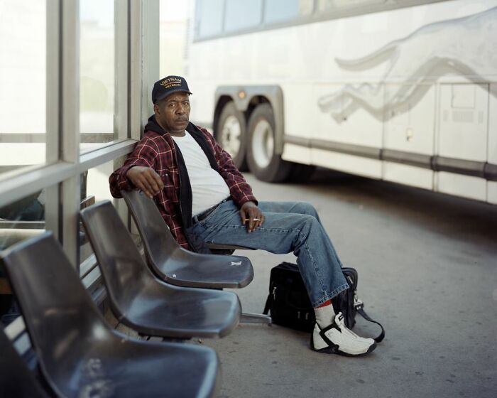 Man wearing a Vietnam veteran hat sitting at a bus stop with a bus in the background, illustrating life at America’s bus stops.