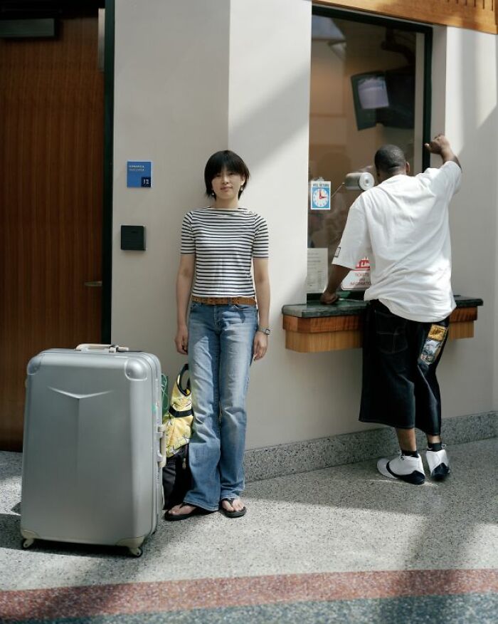 Young woman standing with suitcase at bus station while another person buys ticket, capturing life at America's bus stops.