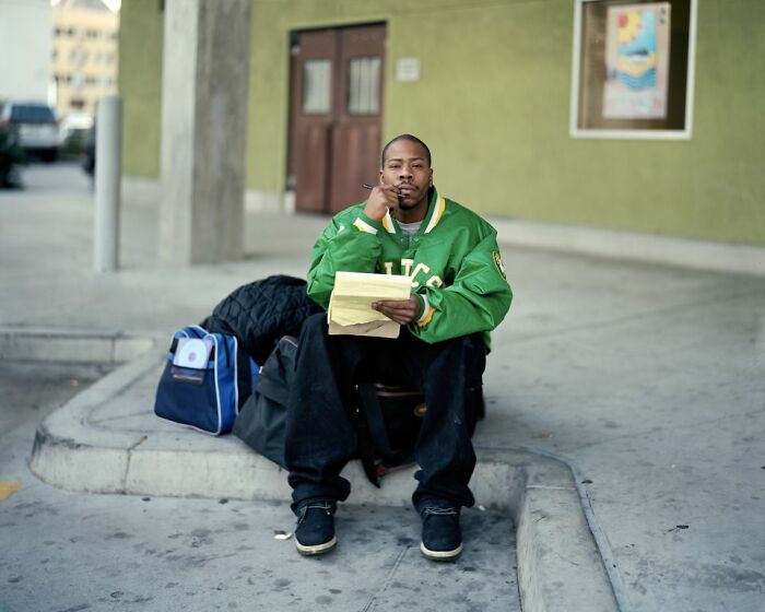 Man wearing green jacket sitting at an America bus stop holding papers with bags beside him on the sidewalk.