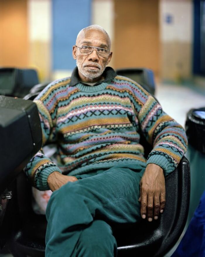 Elderly man sitting at a bus stop, wearing glasses and a colorful sweater, captured in life at America’s bus stops.
