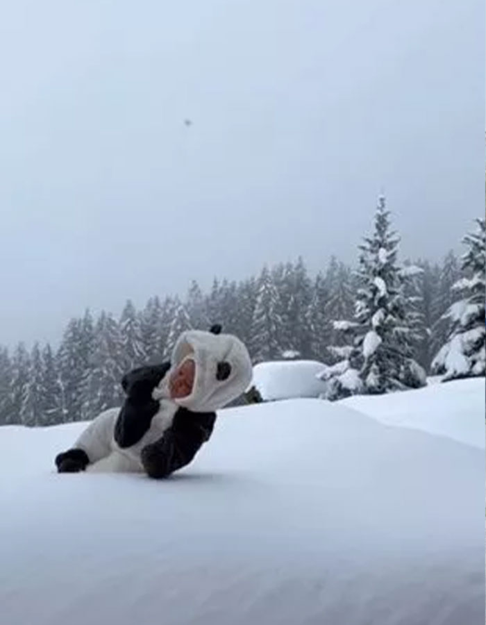 Baby in a panda outfit sitting in a deep snowdrift with snowy trees in the background.