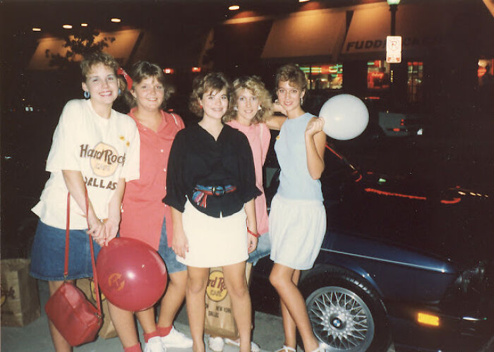 Group of women in 80s fashion trends, wearing denim skirts and colorful tops, holding balloons, standing by a parked car at night.