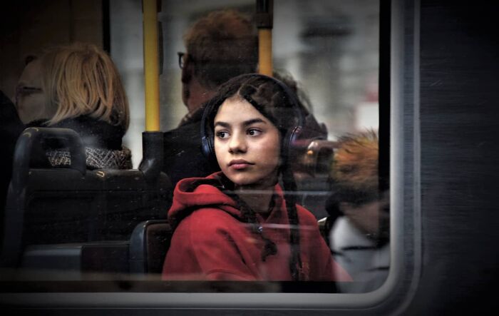 Girl in a red hoodie and headphones on a bus, captured by Nadia Eeckhout, looking out the window at city scenes.