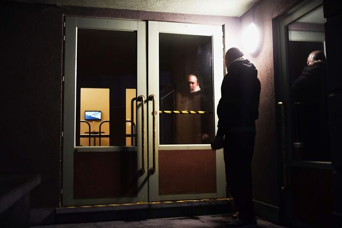 Man standing at a glass door, reflecting the indoors, captured through Nadia Eeckhout's lens depicting everyday moments.