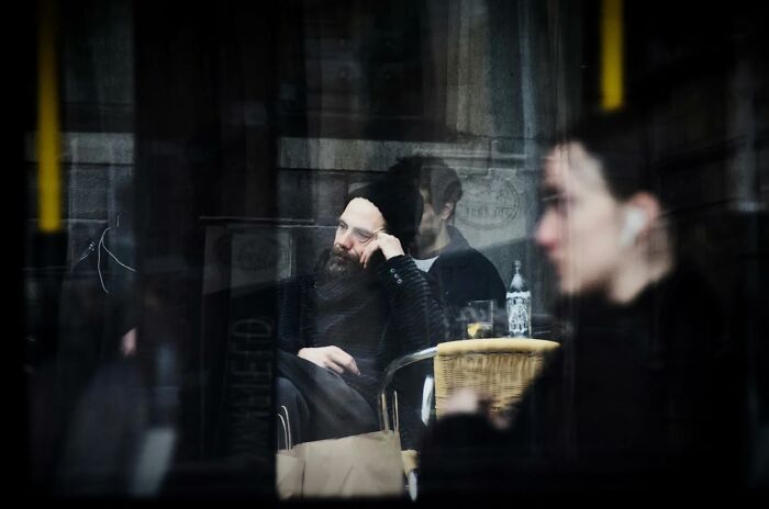 Candid moment captured by Nadia Eeckhout, featuring a man sitting thoughtfully at a cafe reflected in a window.