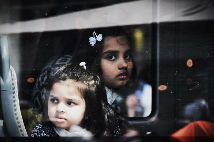 Children sitting on a bus, captured through glass, reflecting everyday moments by Nadia Eeckhout.