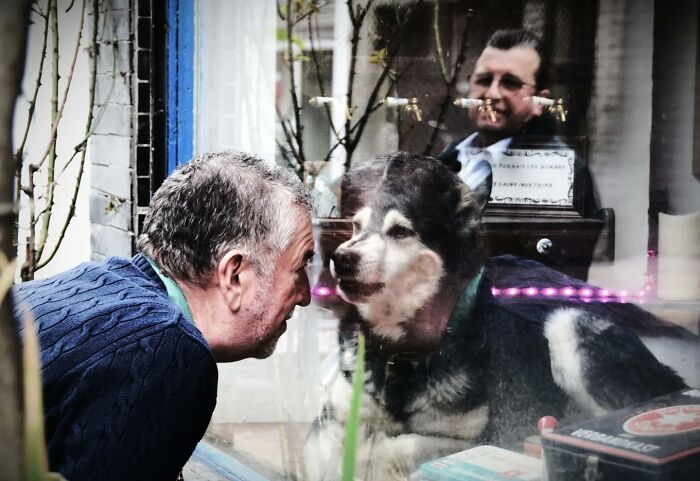 Man and dog share moment through glass, captured by Nadia Eeckhout, with a reflection of another person in the background.