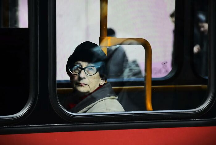 Woman with glasses and hat sitting on a bus, captured in an everyday moment through Nadia Eeckhout's lens.