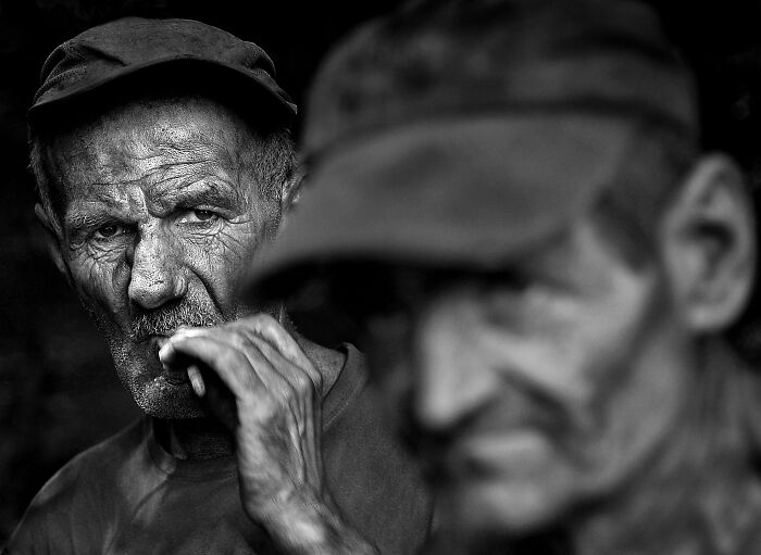 Two men in hats, one in focus with intense gaze, capturing the resilience and beauty of everyday people in black and white.