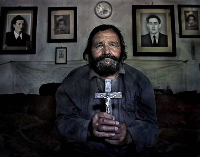 A man holding a crucifix sits in a room with family portraits, capturing resilience and beauty of everyday people.