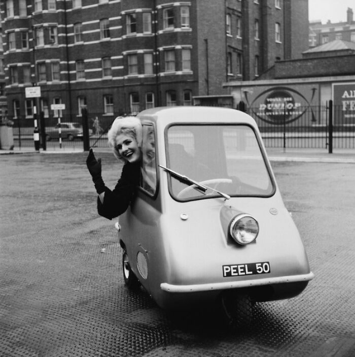 Person driving a Peel P50, a historical invention, in a city street, waving cheerfully from the small car window.