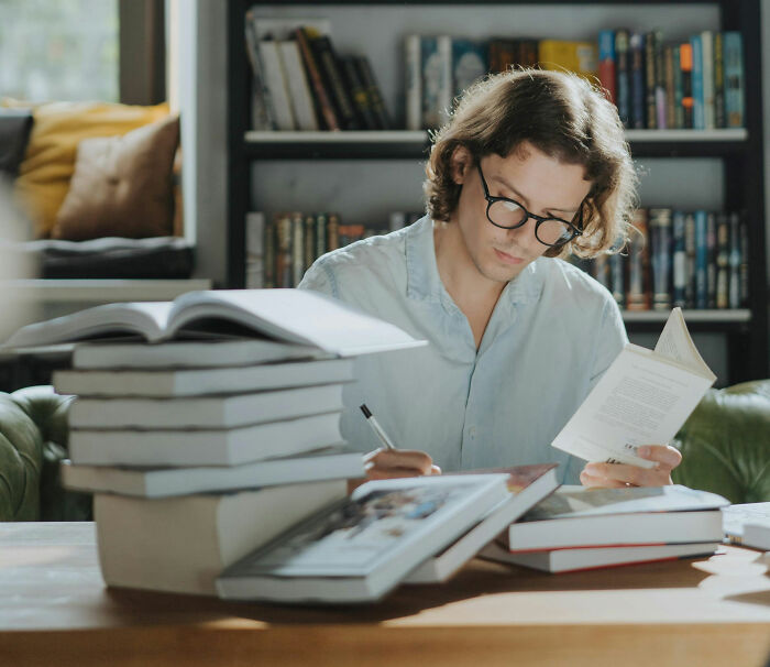 Young man with glasses reading and writing, surrounded by books, portraying mature behavior in a library setting.