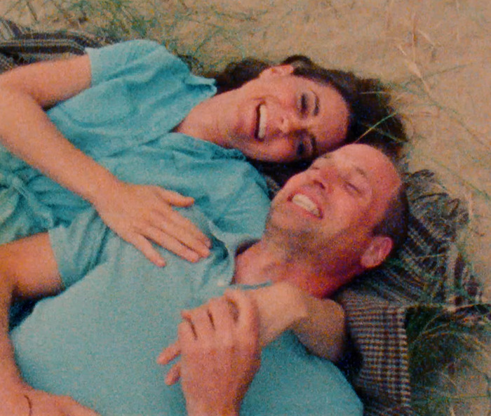 Kate Middleton and Prince William in blue outfits laughing together on a picnic blanket outside.