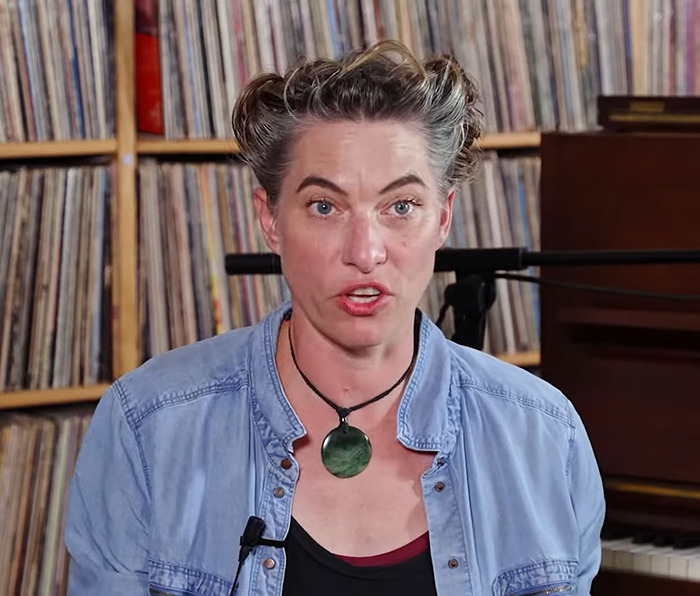 Amanda Palmer sitting in front of shelves filled with records, speaking, and wearing a blue denim shirt and necklace.