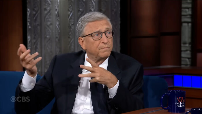 A person gesturing during a talk show segment, wearing a suit, with a mug on the table.
