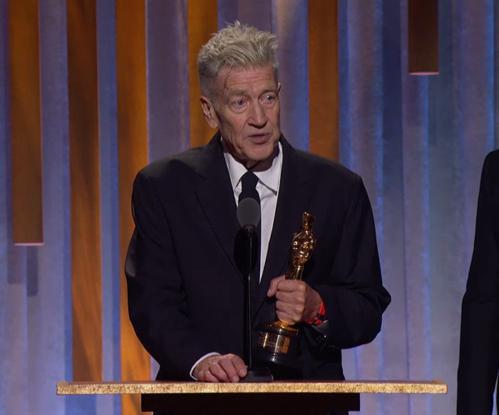 Director David Lynch holding an award, wearing a black suit, in front of a curtain backdrop.