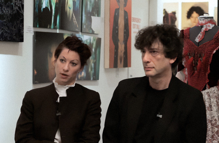 Amanda Palmer and Neil Gaiman sitting in an art gallery, dressed in dark clothing, surrounded by paintings.