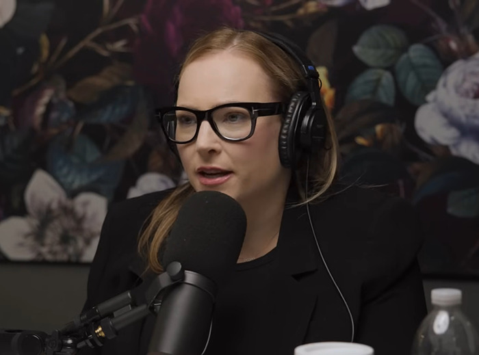 Meghan McCain with glasses and headphones speaking into a microphone, floral background.