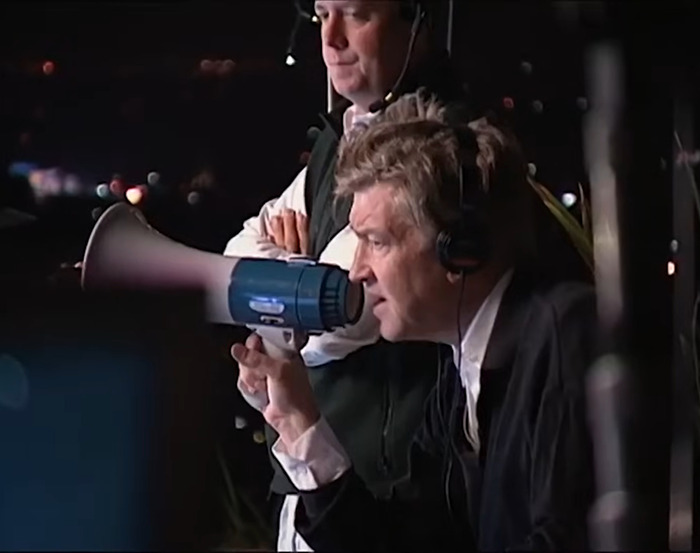 David Lynch using a megaphone on set, wearing headphones, involved in production work.