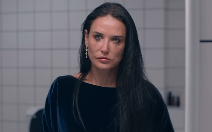 Demi Moore in a blue blouse stands in a tiled room, looking serious.