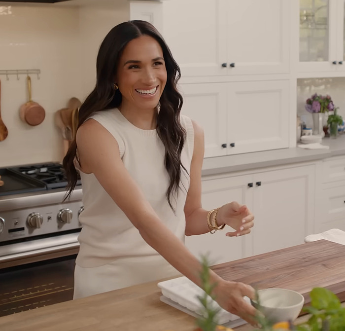 Meghan Markle smiling in a modern kitchen, reaching for a bowl on a countertop, embodying lifestyle themes.