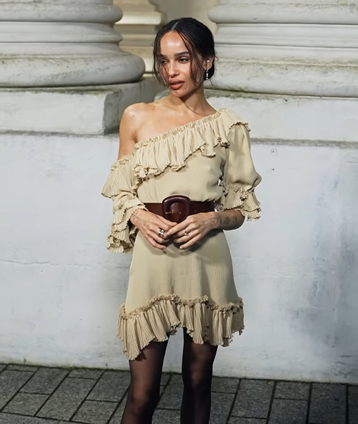Zo&euml; Kravitz in a beige dress, brown belt, and black stockings posing for a photo.