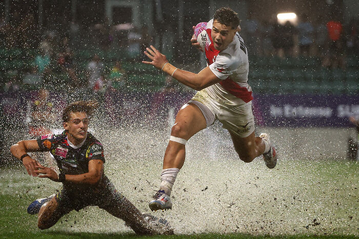 Rugby player leaps over another in dramatic World Sports Photography capture, splashing water into the air.