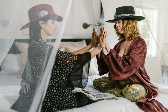 Two women in witchy attire, sitting on a bed, interacting and exploring their witchy personality together.