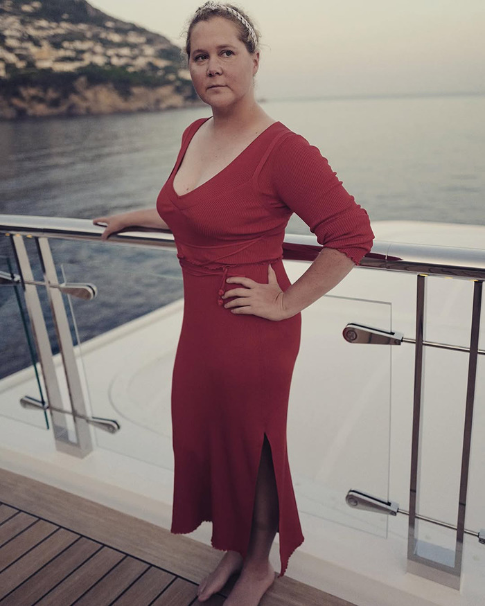 Amy Schumer in a red dress stands on a boat deck by the sea, looking thoughtful.