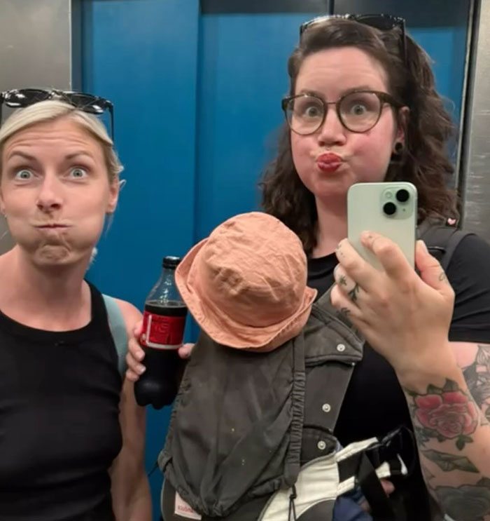 Two moms in an elevator making funny faces, one holding a soda bottle, redefining partying post-baby.