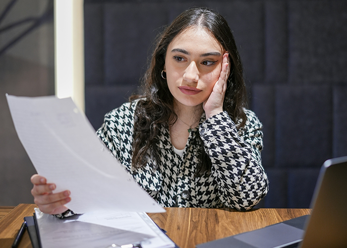 Woman contemplating two weeks' notice at a desk, holding a paper, looking stressed.