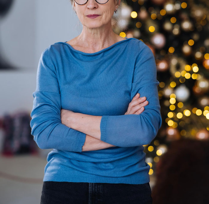Woman in blue sweater with folded arms, standing by a decorated Christmas tree, conveying a tense moment in marriage.