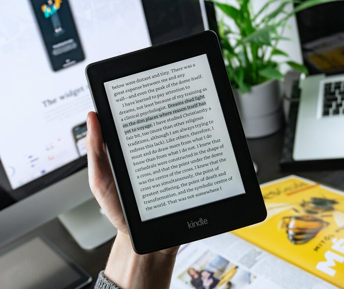 A hand holding a Kindle with text shown on the screen in a modern office setup.