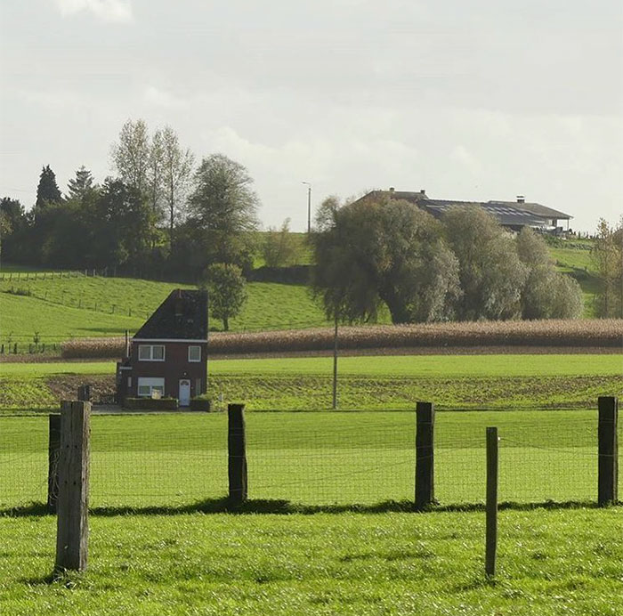 Small, humorously bad house in a vast Belgian landscape, surrounded by green fields and trees.