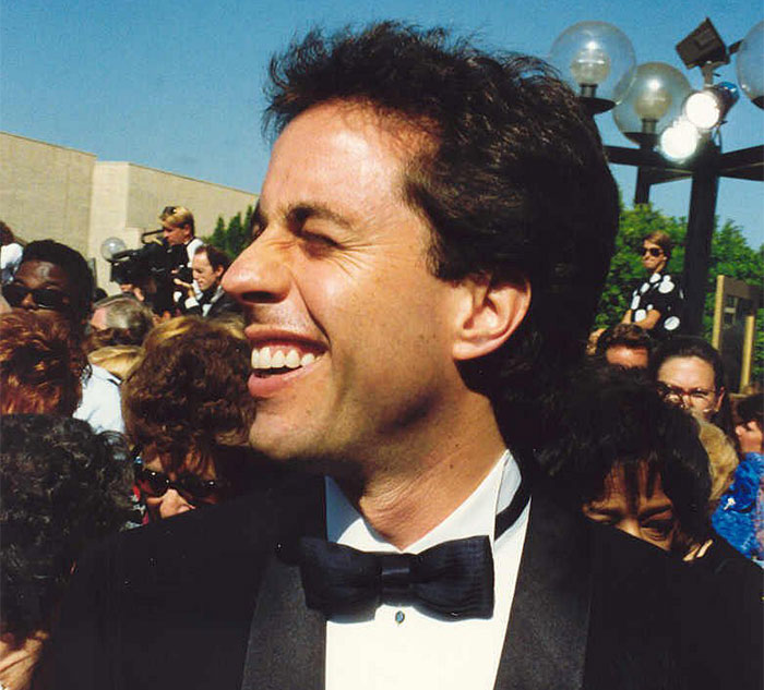 Person in a tuxedo smiling at an outdoor event, surrounded by people and cameras, linked to celebrity secrets and scandals.