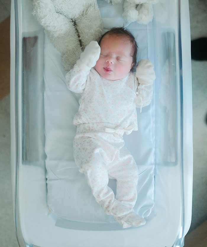 Newborn baby sleeping in a hospital bassinet, wearing a patterned onesie and mittens.