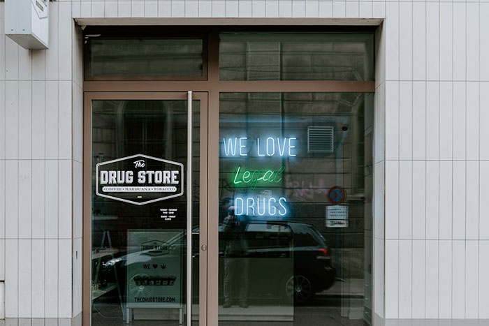 Glass storefront with "The D**g Store" logo and a neon sign saying "We love legal d***s," reflecting car and street.
