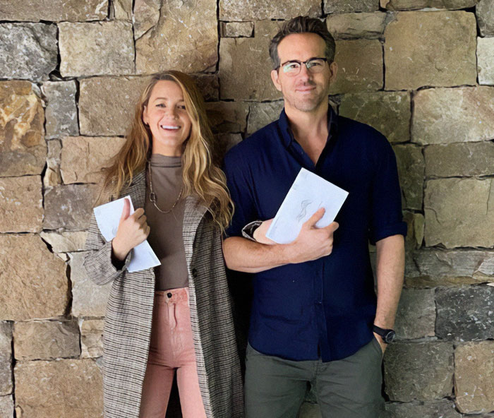 Blake Lively smiling next to a man, both holding papers, posing against a stone wall.