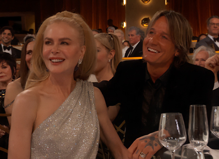 Nicole Kidman in a silver dress at a formal event, sitting with husband Keith Urban, amid other attendees.