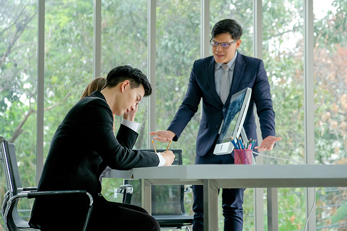 Boss in a suit discussing with an employee at a desk in a modern office setting.